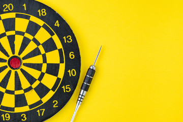 Flat lay of perfect black darts with dartboard on solid yellow background with copy space using as...