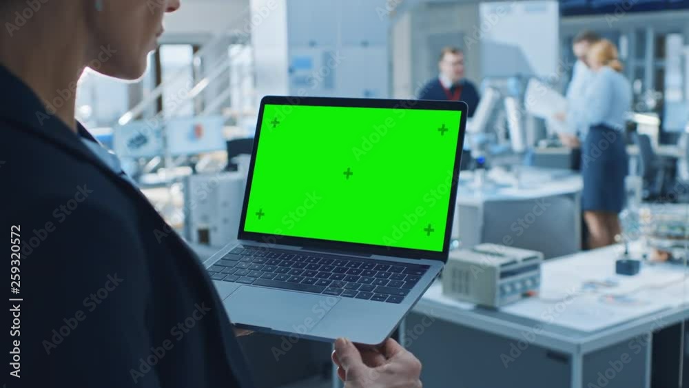 Poster Engineer Holds Green Mock-up Screen Laptop Computer. In the Background Team of Industrial Engineers Working with Functional Robot Arm in Modern Bright Research Facility