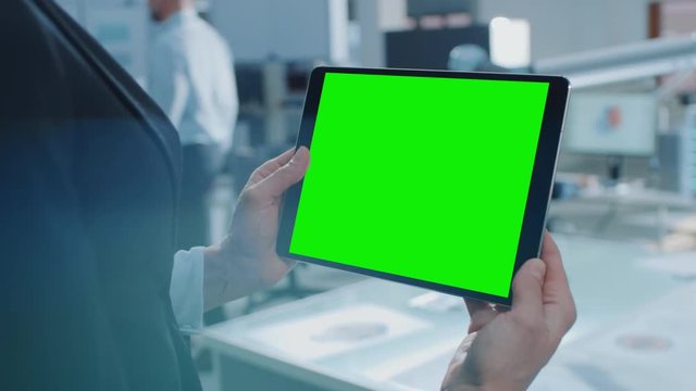 Engineer Holds Green Mock-up Screen Digital Tablet Computer. In the Background Team of Industrial Engineers Working with Functional Robot Arm in Modern Bright Research Facility