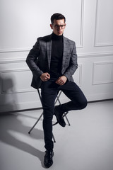 Strong,stylish man in elegantly costume ,black eyeglasses sit on black chair and look serious in one direction.Studio photoshoot on the white wall.Vertical view