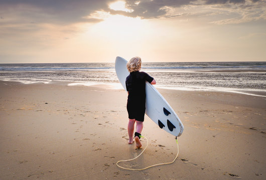 Blonder Junge läuft am Strand mit Surfbrett Richtung Meer bei Sonnenuntergang
