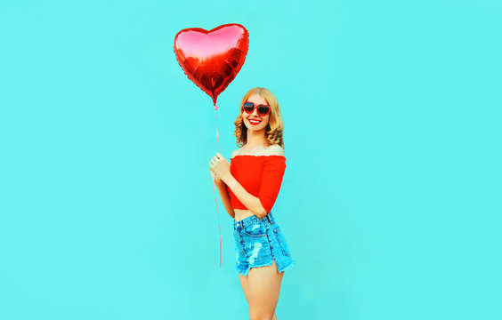 Portrait cute happy smiling woman in shorts holding red heart shaped air balloon on colorful blue background