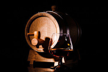 Glass of cognac with oak barrel on black background. Wooden barrel for whiskey with glass.