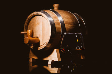 Glass of cognac with oak barrel on black background. Wooden barrel for whiskey with glass.