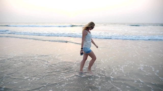 A playful young long-haired blonde girl walks along the beach on the water of the sea jumps up and creates splashes of feet and waves her hand greeting someone. Happy woman resting on the sea vacation