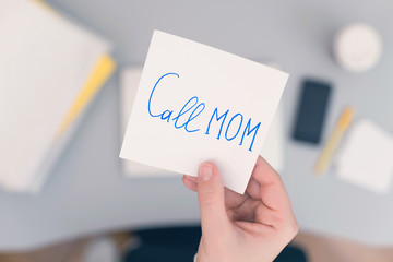 Woman clerk is sitting at office table holding note sticker with message "call mom" and going to call up her mother.