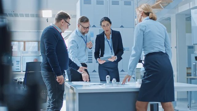 Diverse Team of Industrial Robotics Engineers Gathered Around Table With Robot Arm, They Use Tablet Computer to Manipulate and Program it to Pick Up and Move Metal Component. Bright Facility
