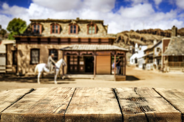 Naklejka premium Wooden old table of free space for your decoration and wild west blurred background of big building and cowboy on horse. 