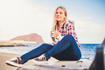 Fototapeta na wymiar Cheerful beautiful blonde tourist sit down on her black car and enjoy the outdoor activity feeling the nature - young traveler people with ocean and mountain in background