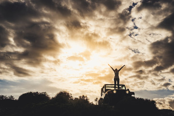 Fototapeta premium Success and lonely traveler concept with girl in silhouette standing on his car and opening arms up to the dramatic sky of a golden sunset - outdoor traveler adventure with vehicle