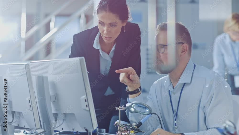 Poster female chief engineer talks with electronics specialist, explaining things, he works on computer and