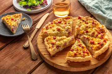 A photo of a quiche with a piece cut off, with wine and salad leaves on a dark rustic wooden...