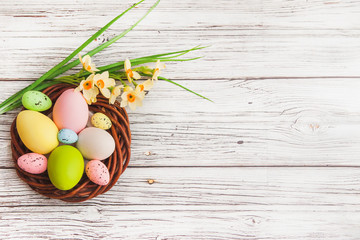 Happy Easter. Congratulatory easter background with copy space. Colorful Easter eggs in the nest and narcissus flowers on light wooden background
