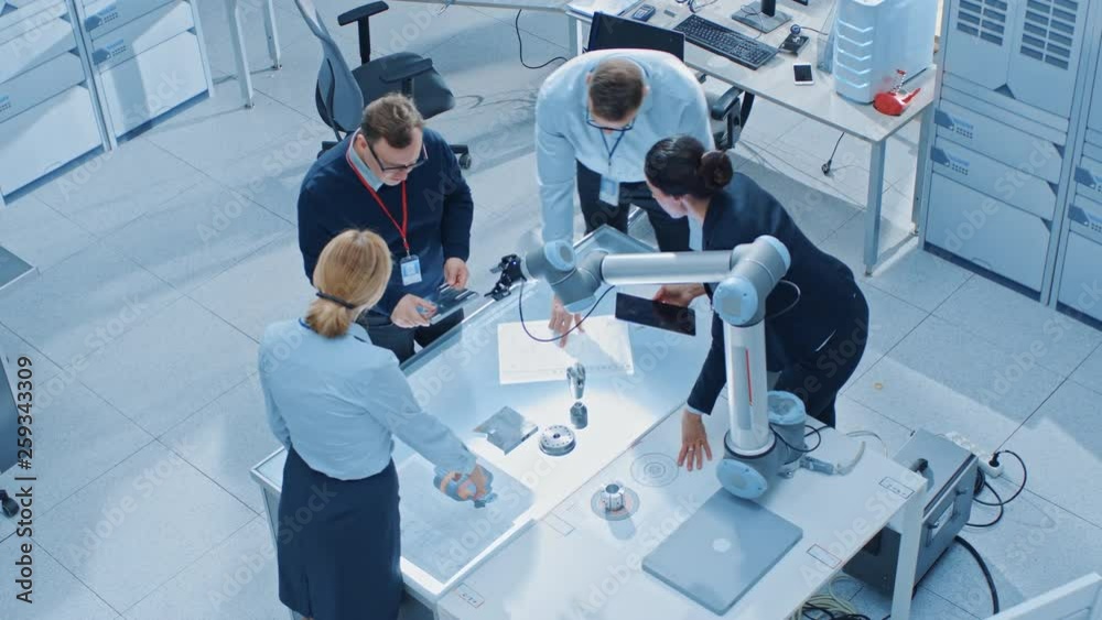 Leinwandbilder Team of Industrial Robotics Engineers Gathered Around Table With Robot Arm, They Use Tablet Computer to Manipulate and Program it to Pick Up and Move Metal Component. Elevated Above Shot
