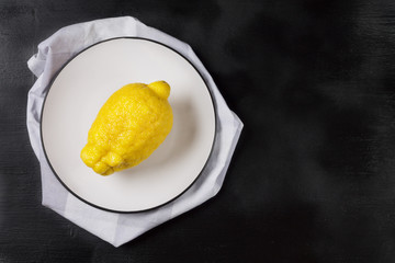 One lemon on a white plate on a light kitchen towel on a black background. Trendly ugly organic fruit. Copy space.
