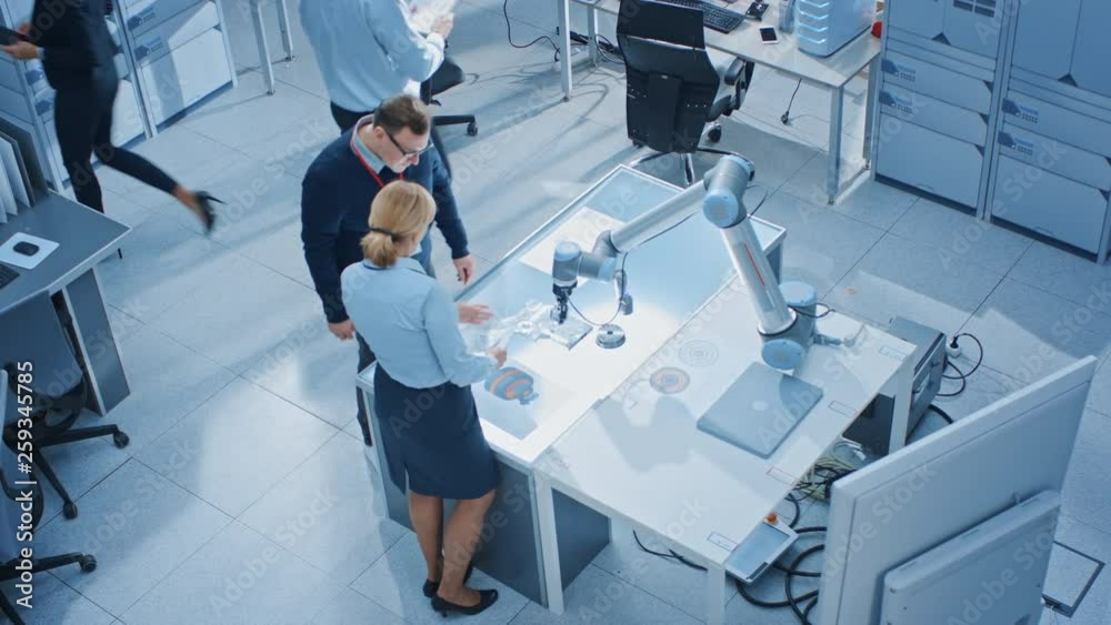 Fototapete Team of Industrial Robotics Engineers Working on Computers, Talking and Using Machine Learning Technique to Manipulate and Program it to Pick Up and Move Metal Component. Top Down Above Shot
