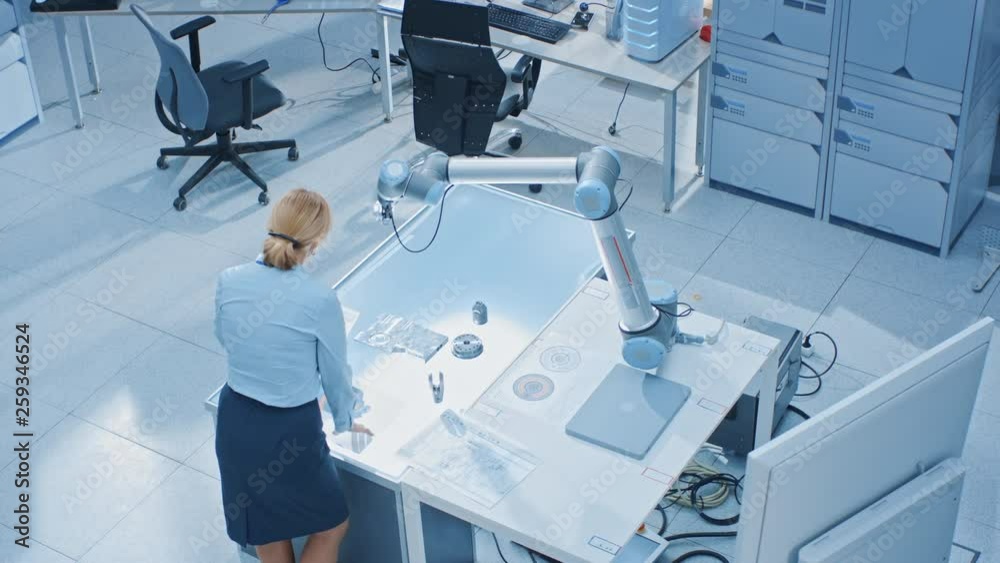 Fototapete Stylish Female Industrial Robotics Engineer Looks at Blueprints Engineering Ways to Program and Manipulate Robot Arm for Lifting, Moving Objects. Technology Research Facility for Cloud Computing