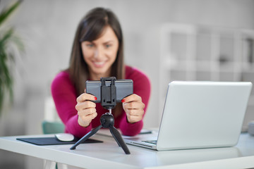 Charming Caucasian brunette dressed casual adjusting smart phone for video call. In front of her laptop, office interior.