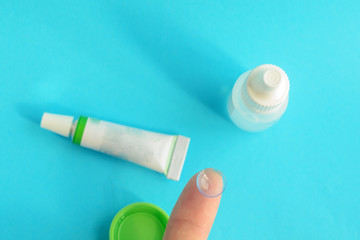 Contact lens on finger, tube of eye ointment and small bottle of eye drops on light blue background..Top view.