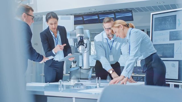 Diverse Team of Industrial Robotics Engineers Uses Tablet Computer to Program and Manipulate Robot Arm for Lifting Moving Objects. Technology Research Facility for Machine Learning and Cloud Computing