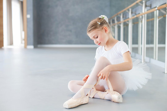 Little ballerina is trying on pointe shoes in ballet class. Cute kid girl is sitting on floor under barre. Child is wearing in white ballet clothes and dancing dress with tutu skirt.