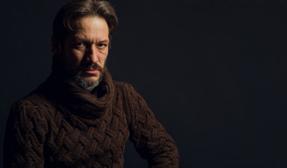 Portrait of a man on a black background. The model sits in a brown knitted sweater. Gray beard and confident look saturated with strength and courage