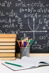 School desk in classroom, with books on background of chalk board with written formulas. Soncept Teacher's Day.