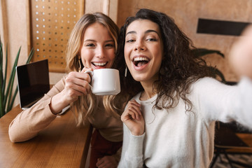 Excited happy pretty girls friends sitting in cafe take a selfie by camera.