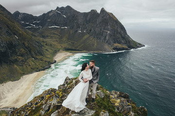 Naklejka premium Wedding couple travelers on a hill in Norway, Kvalvika. Beautiful view of the beach, Lofoten, Norway.