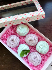 Homemade marshmallows of different colors are beautifully packed in a gift box. Decorated with mint leaves. Nearby is the lid of the box with a transparent window. On a dark background.