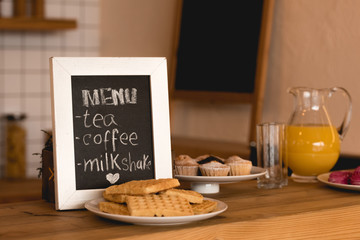 menu board, dishes with waffles and cakes, and jug with fresh juice on bar counter in coffee shop