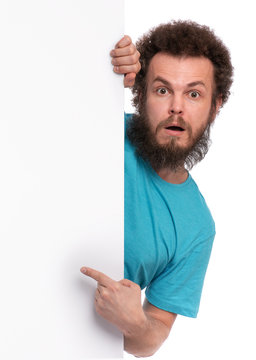 Crazy Bearded Man With Funny Curly Hair Showing Empty Blank Signboard With Copy Space. Guy With Surprised Eyes And Mouth Open Peeking Out From Behind Big White Banner, Isolated On White Background.