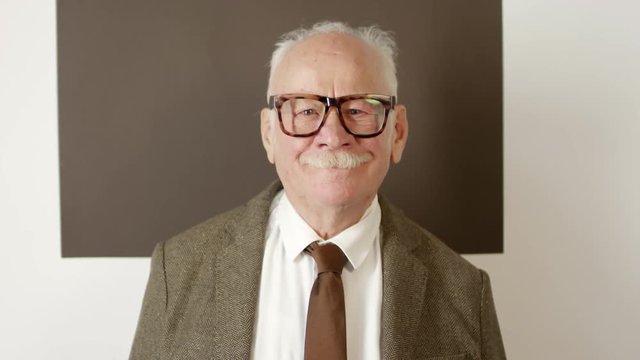 Portrait of elderly man wearing tweed jacket, necktie, and glasses and leopard pattern standing against white and brown background and looking away, then turning towards camera and smiling