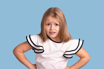Close up emotional portrait of young blonde  girl  on blue background in studio. She  holds her hands at the waist and is  complaining about something  or expresses dissatisfaction with something.
