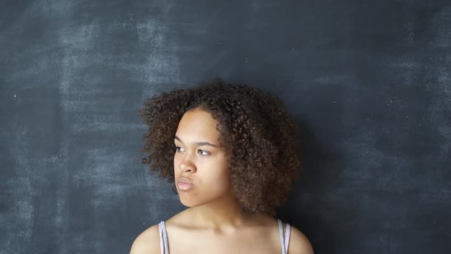 Tilt down portrait shot of serious young black woman with curly hair standing against dark chalkboard wall and looking at camera