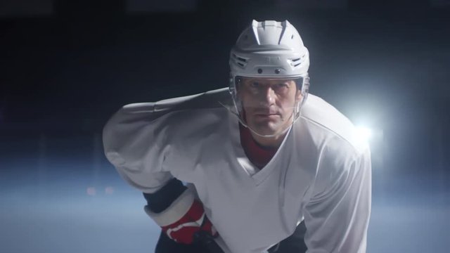 Portrait of determined middle-aged Caucasian ice hockey player in helmet, jersey and gloves standing on rink and looking at camera, medium shot