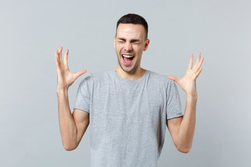 Portrait of crazy screaming young man in casual clothes keeping eyes closed, spreading hands isolated on grey wall background in studio. People sincere emotions, lifestyle concept. Mock up copy space.