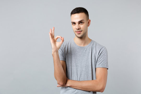Portrait of handsome confident young man in casual clothes standing, looking camera showing OK gesture isolated on grey wall background. People sincere emotions, lifestyle concept. Mock up copy space.