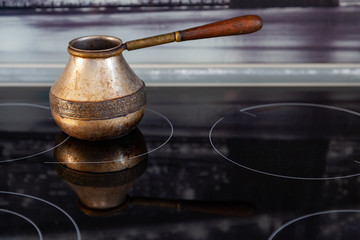 Old Bronze Turkish retro coffee maker kanaka on glass hob and stove with wooden handle, worn on a black background in an apartment in the kitchen. Retro cezve, briki, rakweh, pot on a stovetop.