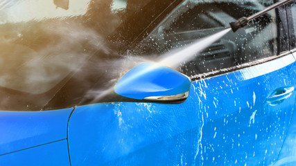 Side mirror of blue car washed in self serve carwash, jet water sprays from high pressure nozzle.