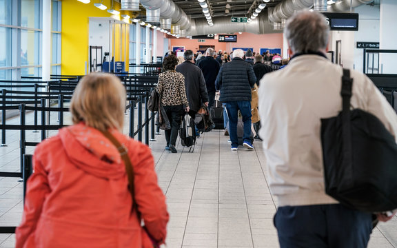 Group Of Anonymous People At Airport Gate Line To Board An Airplane, Queue Crowd, Pulling Their Trolley Seen From Behind.