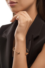Cropped half-turn portrait of young lady with tanned skin, wearing black sleeveless blazer. The girl with slight smile is wearing golden open bracelet, adorned with triangle scroll elements on edges.