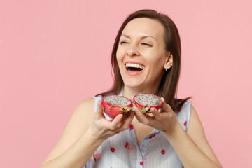 Laughing young woman looking aside, holding halfs of fresh ripe pitahaya, dragon fruit isolated on pink pastel background in studio. People vivid lifestyle relax vacation concept. Mock up copy space.