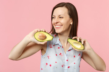 Smiling young woman in summer clothes looking aside, hold halfs of fresh ripe avocado fruit isolated on pink pastel wall background. People vivid lifestyle relax vacation concept. Mock up copy space.