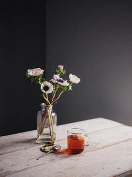 Eine Glastasse mit Tee neben Blumenvase mit Anemonen und Zitrone auf einem Keramikteller