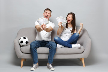 Overjoyed couple woman man football fans cheer up support favorite team holding fan of money in dollar banknotes cash money doing winner gesture isolated on grey wall background. Sport family concept.