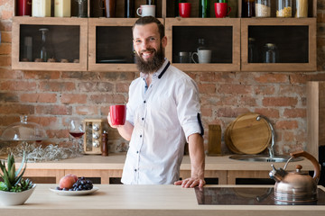 Hot morning drink. Energy and vitality. New day. Young man with red cup smiling looking at camera. Loft kitchen space.