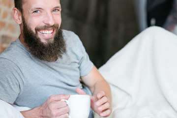 Morning drink warmth and energy. New day. Rise and shine. Smiling hipster in bed with cup of beverage.