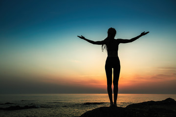 Fototapeta premium Silhouette of a flexible yoga woman stands on the shore of the ocean at evening.
