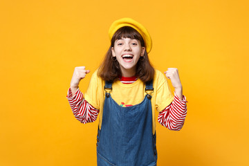 Portrait of cheerful girl teenager in french beret, denim sundress clenching fists like winner isolated on yellow background in studio. People sincere emotions, lifestyle concept. Mock up copy space.